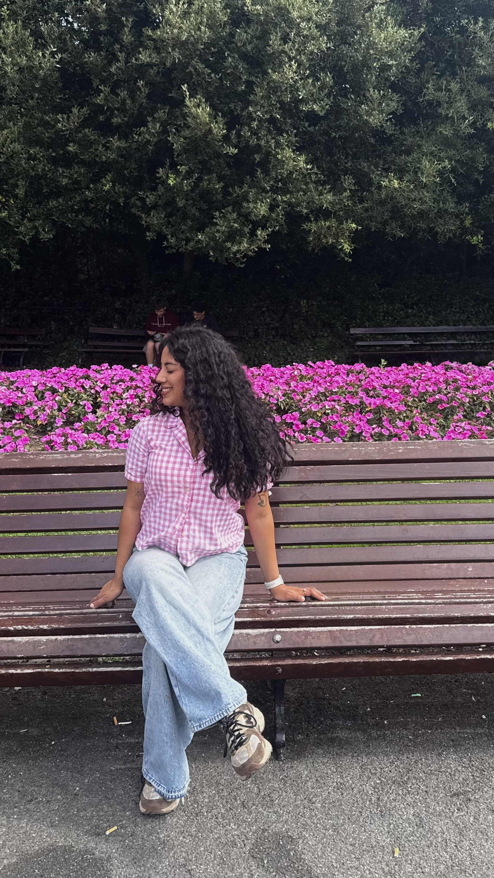 A woman sitting on a wooden bench in front of a vibrant flower bed filled with pink blossoms. Behind the flowers is a dense row of green trees. The woman is wearing a pink checkered short-sleeve shirt, light blue jeans, and sneakers, with long curly hair cascading over one shoulder.
