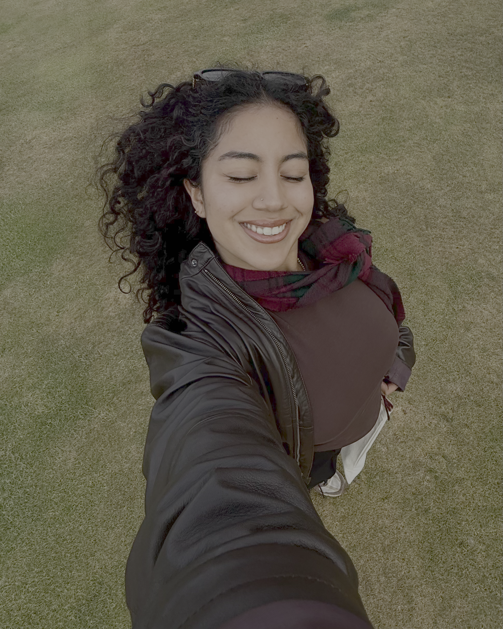 A woman is taking a selfie from an overhead angle while standing on a grassy field. She is wearing a dark leather jacket over a brown top, a multicolored scarf, and sunglasses resting on her head. Her arm is extended outward holding the camera.
